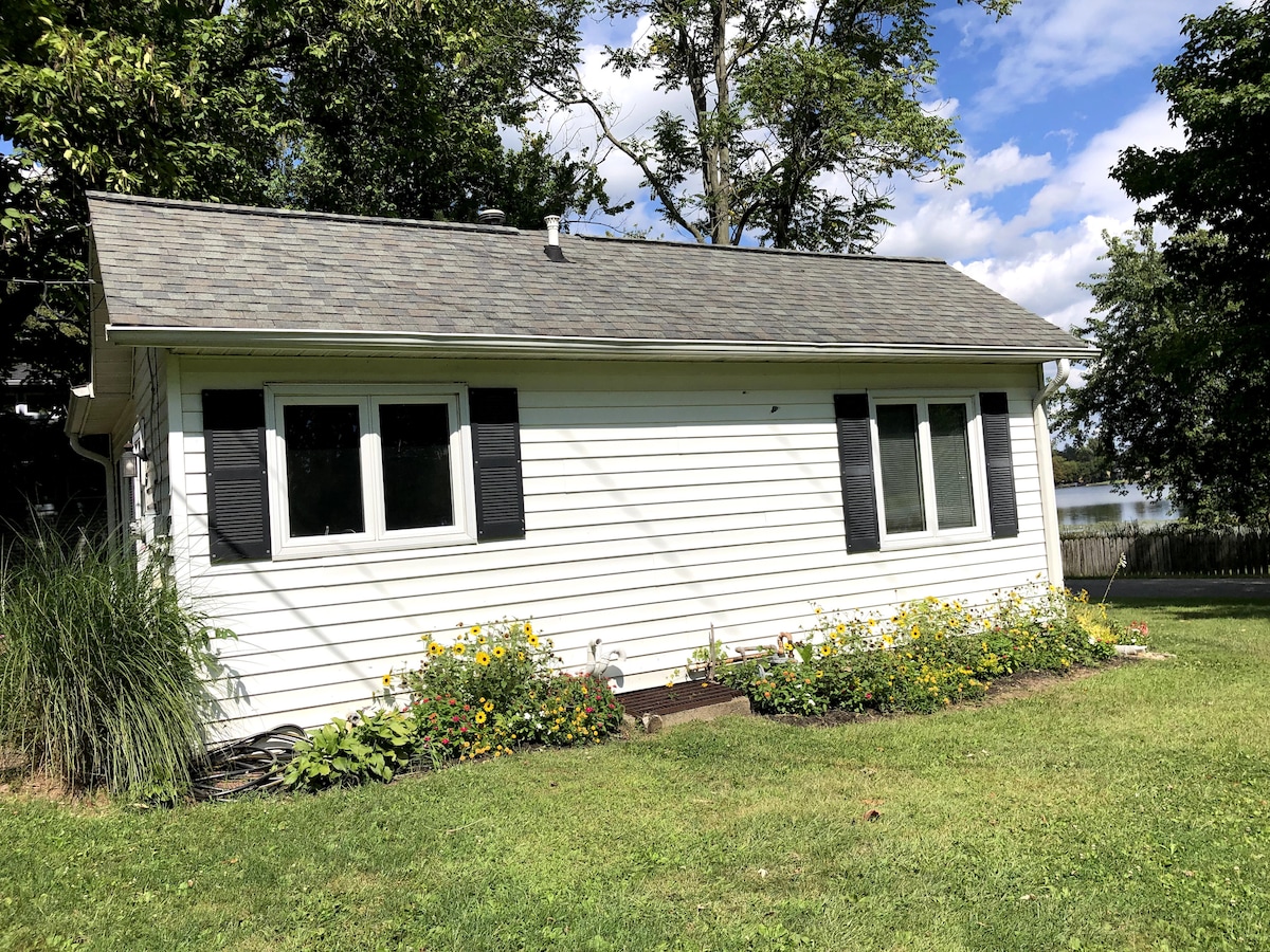 A charming cottage exterior is surrounded by lush greenery and colorful flower beds. The white siding is contrasted by black shutters on the windows. A view of the lake is visible in the background, under a partly cloudy sky.