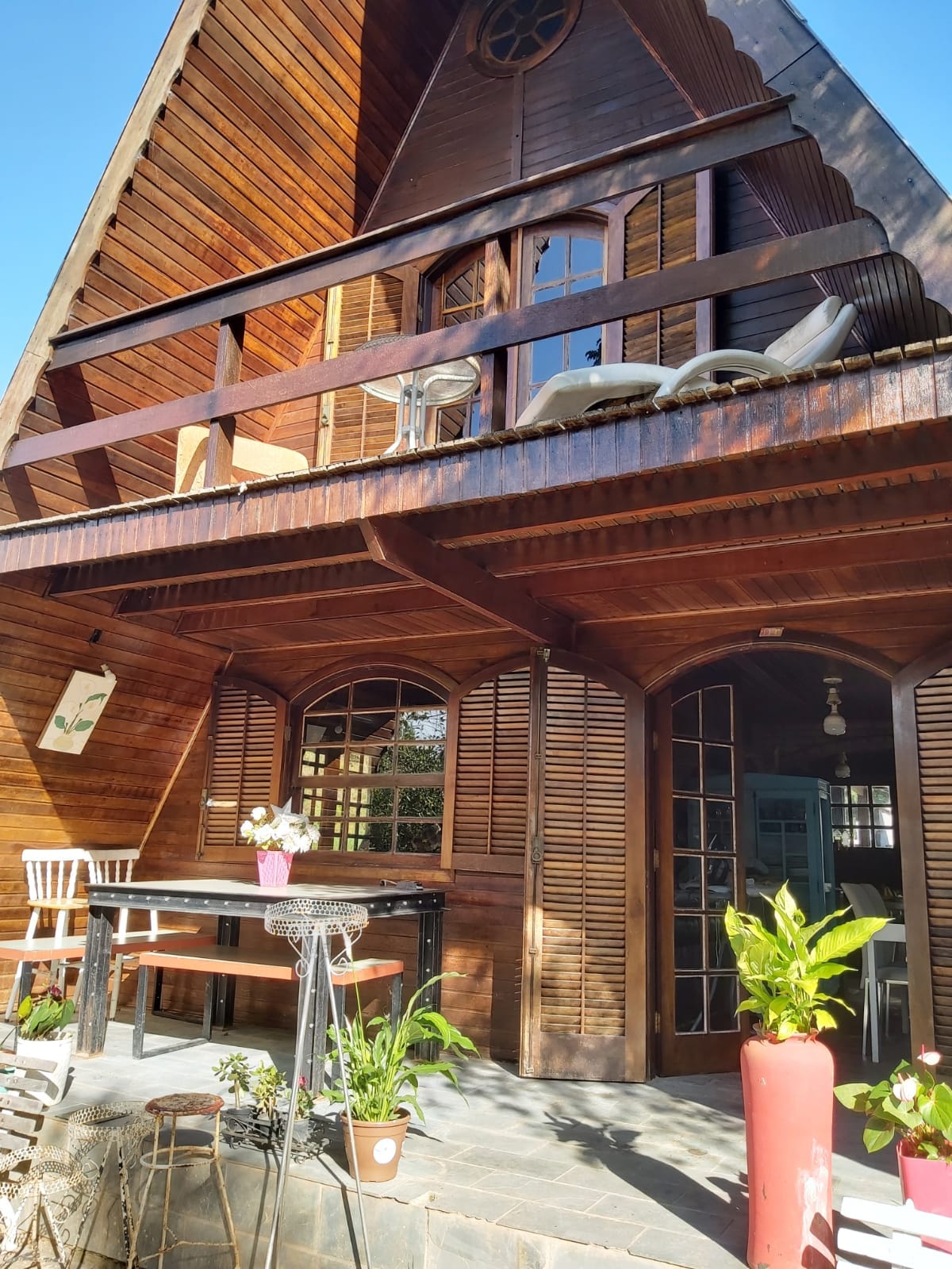 A charming wooden chalet features an A-frame design with a covered balcony. An outdoor seating arrangement includes a table decorated with a vase of flowers. Potted plants are positioned near the entrance, enhancing the inviting appearance of the space.