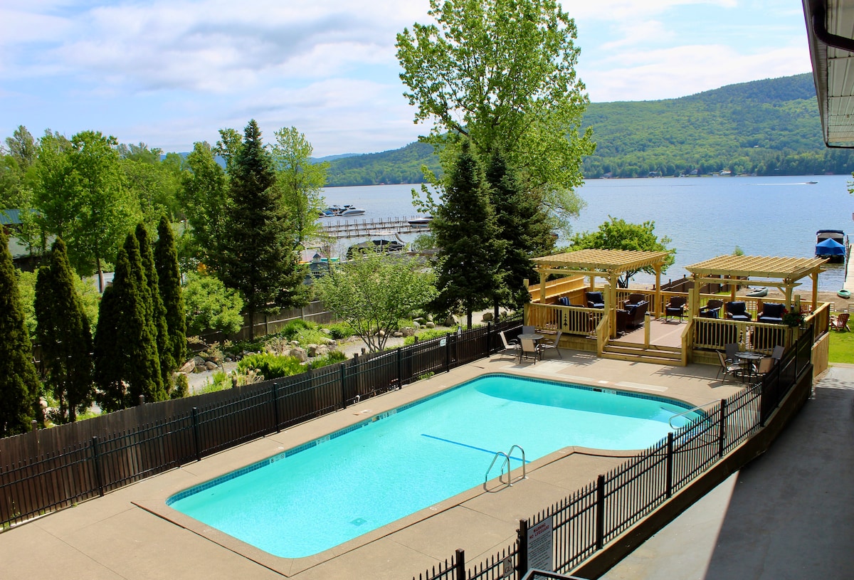 A sunny outdoor area features a clear blue pool surrounded by a gated concrete deck. Lush greenery, including trees and shrubs, creates a natural backdrop, while the calm lake can be seen in the distance, harmonizing with the landscape.
