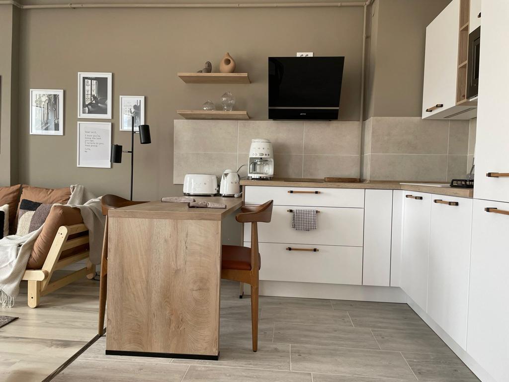 A modern kitchen area is visible, featuring clean white cabinetry and wooden shelving. A small dining table with two chairs complements the space, while a sleek black television hangs above the countertops. Subtle decor items and a coffee maker are arranged on surfaces.