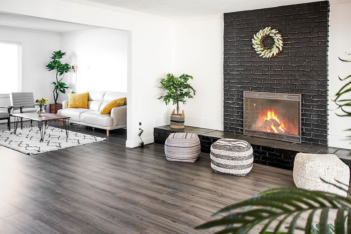 A modern living area features a cozy white sofa accompanied by patterned pillows, with a coffee table placed in front. A fireplace with a decorative wreath stands against a black brick wall. Plants are arranged around the space, providing a touch of greenery.