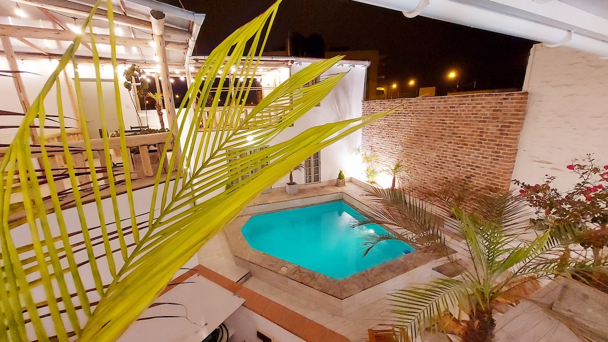 An inviting pool area is framed by lush green palm leaves, with a turquoise pool surrounded by stone decking. The rustic brick wall is softly illuminated, enhancing the serene ambiance. Twinkling lights create a pleasant atmosphere for evening gatherings.