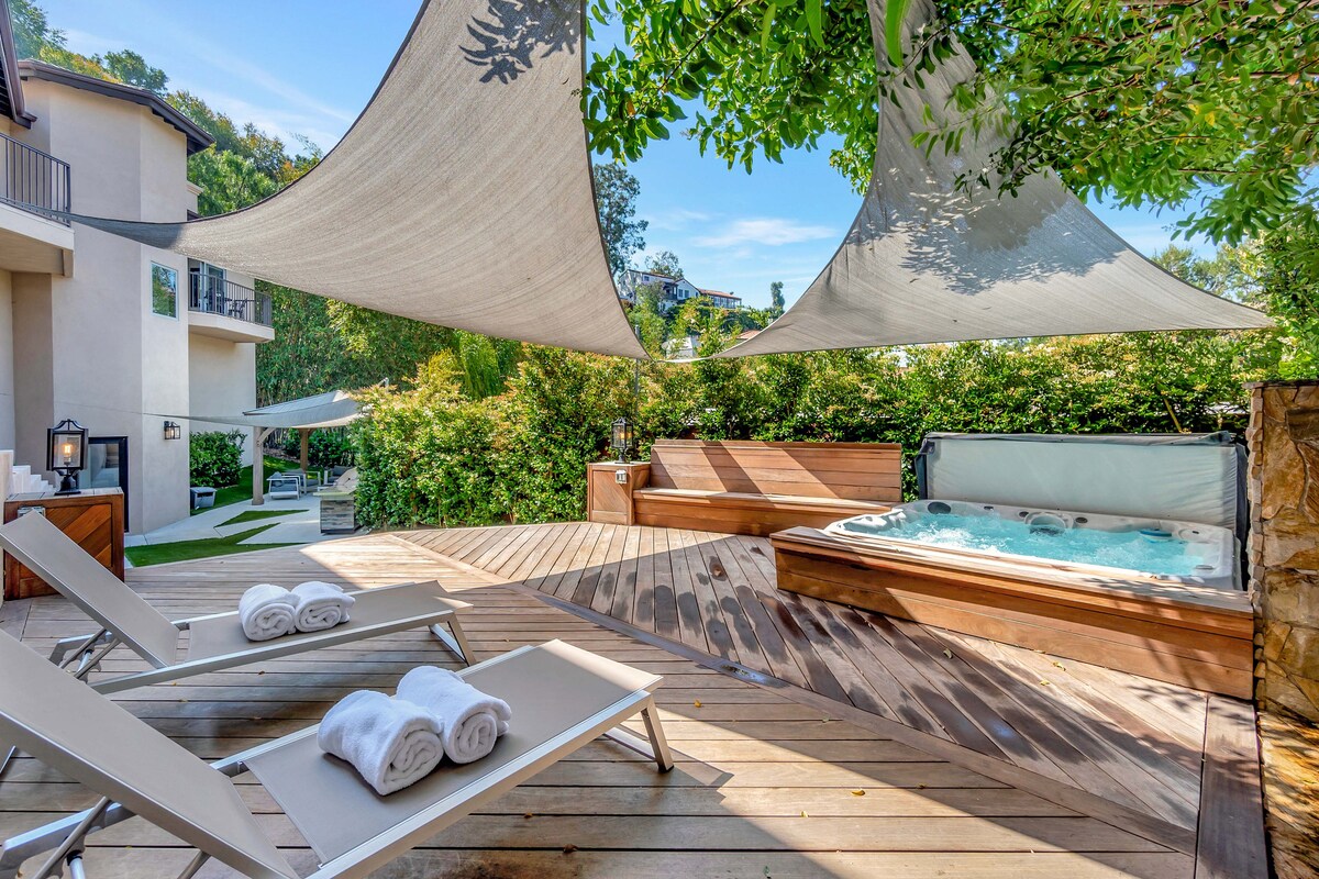 A serene outdoor space is presented with a wooden deck, featuring lounge chairs accompanied by neatly rolled towels. A hot tub is situated nearby, framed by vibrant greenery and shaded by large fabric canopies, creating a peaceful atmosphere for relaxation.