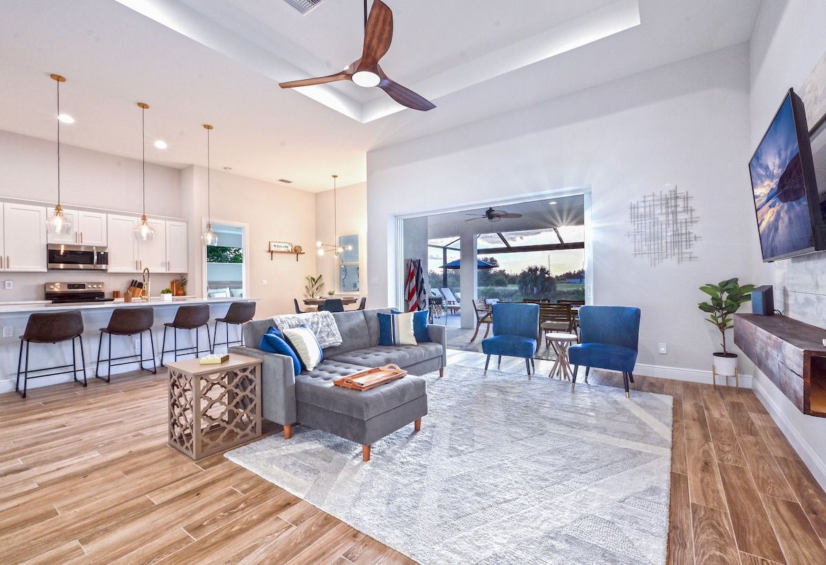 A modern, open-concept living area features a grey sectional sofa accompanied by blue accent chairs. A stylish coffee table sits in front of the seating arrangement, with a large area rug beneath. Large sliding doors provide a seamless transition to the outdoor space, allowing natural light to fill the room.