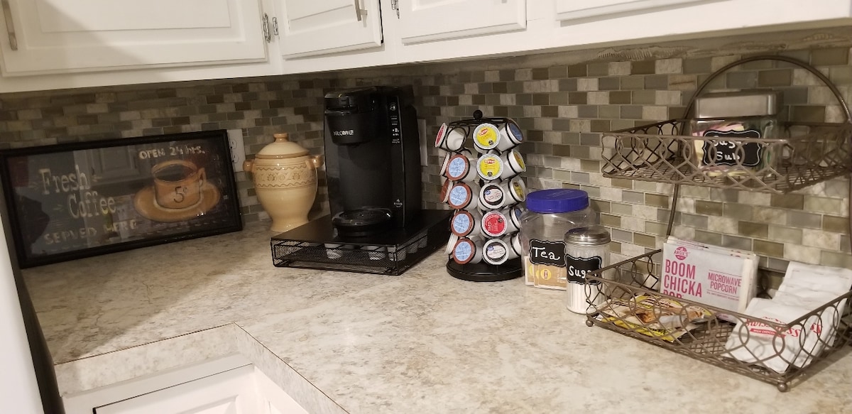 The kitchen counter features a coffee maker, a rotating holder with assorted coffee pods, and a functional wire basket containing tea bags and snacks. A decorative jar and a framed sign reading 'Fresh Coffee Served Here' enhance the space.