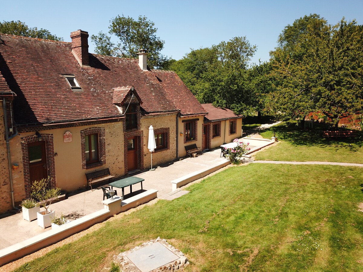 A charming longère is depicted, showcasing its rustic brick façade and inviting outdoor terrace. Sunlight bathes the spacious lawn, framed by trees. Outdoor seating is provided, along with an umbrella for shade, creating a peaceful space for relaxation.