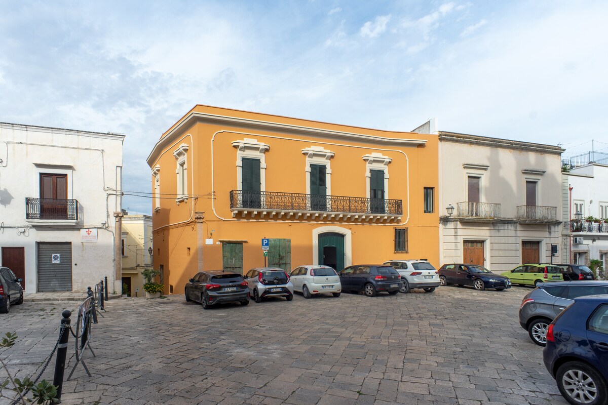 A historic building with a vibrant orange facade is positioned in a lively square. Decorative balconies overlook the street, while surrounding parked cars add to the bustling atmosphere. The architecture reflects classic design elements, contributing to the character of the central area.