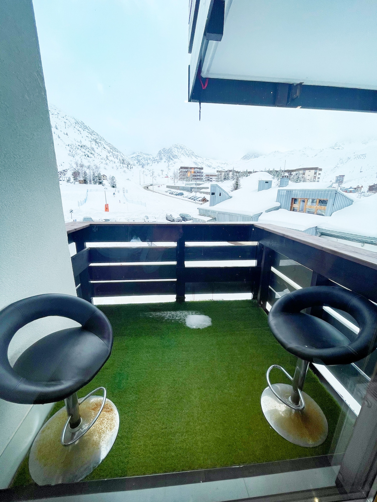 A small balcony features two bar stools and artificial grass flooring. Snow-covered mountains are visible in the background, with distant buildings nestled among the landscape. The balcony provides a relaxed outdoor space with a clear view of the serene winter scenery.