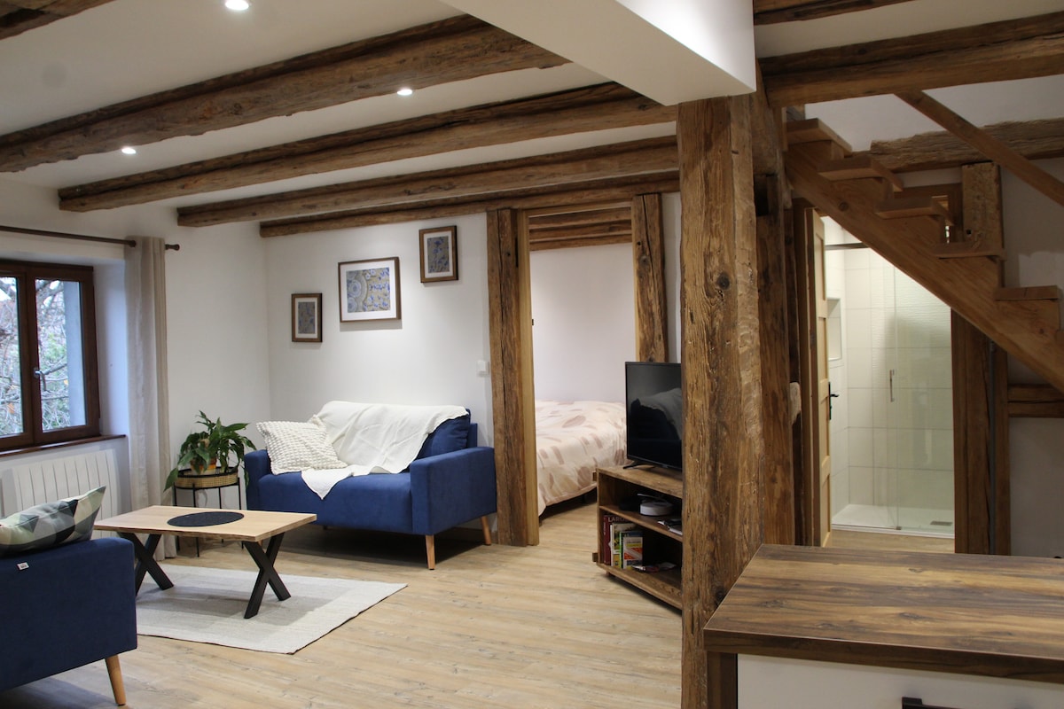 A spacious living area features exposed wooden beams and rustic charm. Two blue sofas are arranged around a round coffee table, with light streaming in through a window. A staircase leads to the upper floor, while a glimpse of a well-lit bedroom is visible.
