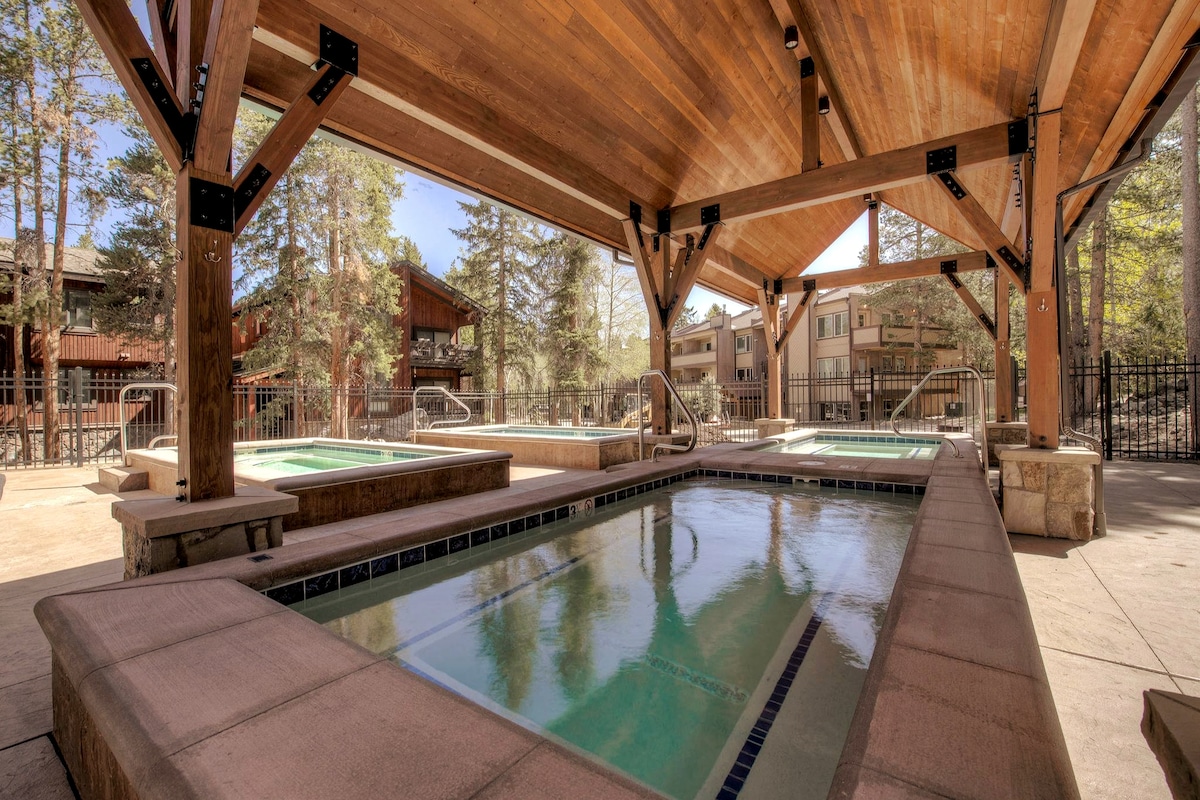 A spacious outdoor area houses multiple hot tubs beneath a large wooden pavilion. The soothing water surfaces reflect surrounding trees, and a peaceful atmosphere is created by the rustic design. A fenced perimeter provides privacy while enhancing the tranquil setting.