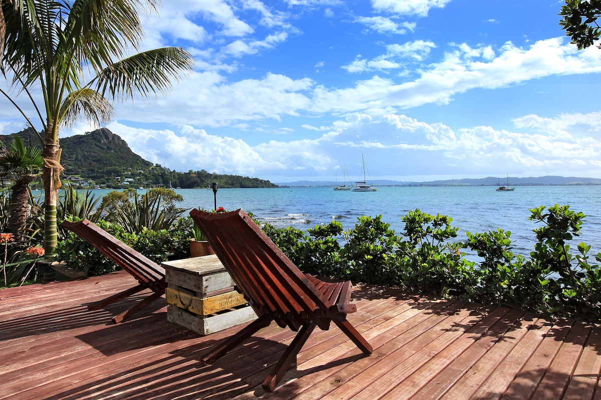 A wooden deck is presented with two angled lounge chairs overlooking a serene bay. Lush greenery surrounds the edge, while views of boats and distant hills enhance the coastal scenery under a partly cloudy sky.