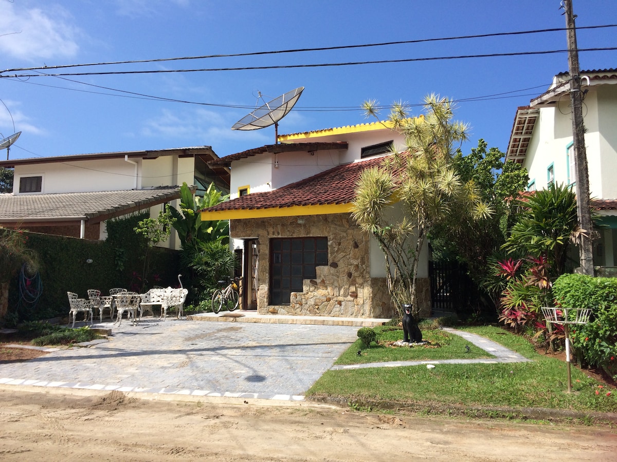 A charming two-story house is depicted, featuring a stone façade and a red-tiled roof. A spacious yard is visible, bordered by lush greenery. Outdoor seating is provided by white metal chairs arranged on a paved area. Satellite dishes are mounted on the roof.
