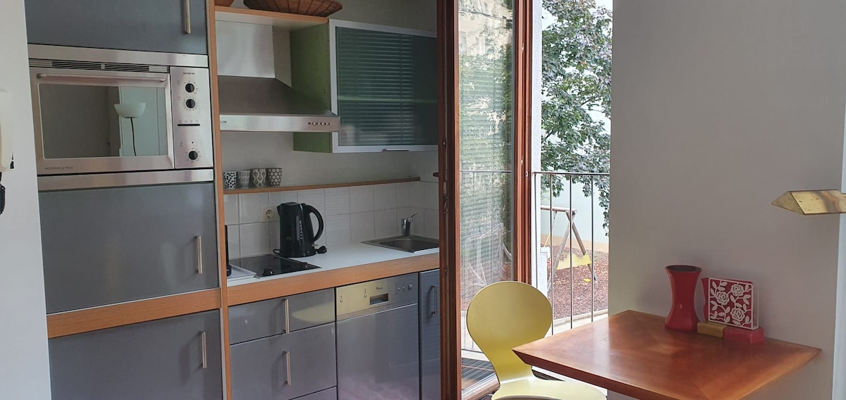 A modern kitchen area is displayed, featuring sleek cabinetry and stainless steel appliances. A compact dining table is positioned near a large window, allowing natural light to fill the space. A small balcony is visible, providing outdoor access.