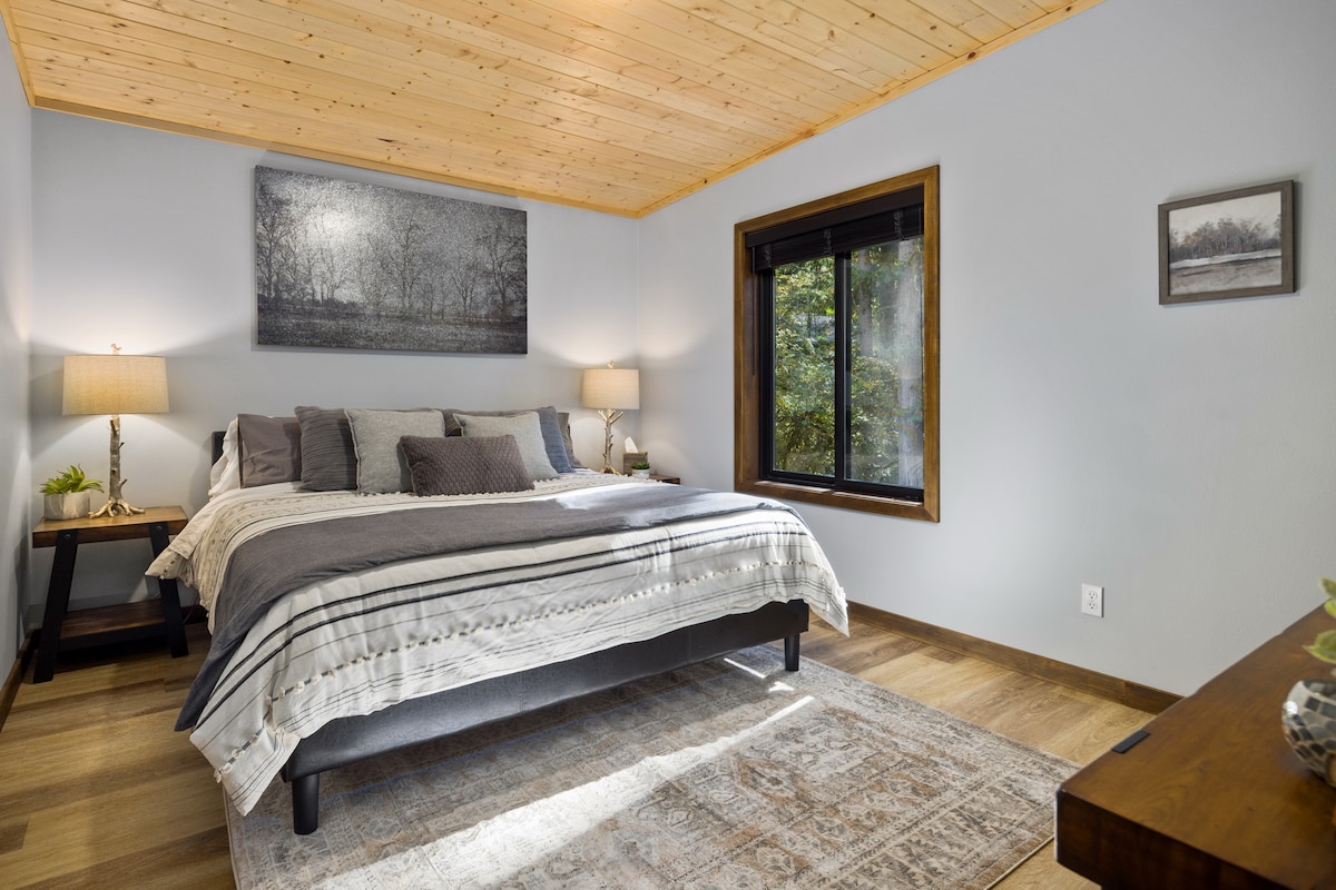 A serene bedroom features a spacious bed layered with neutral-toned bedding and several decorative pillows. Two bedside lamps provide soft light, while a large window invites natural light and offers views of the surrounding trees. A subtle area rug complements the wood flooring.