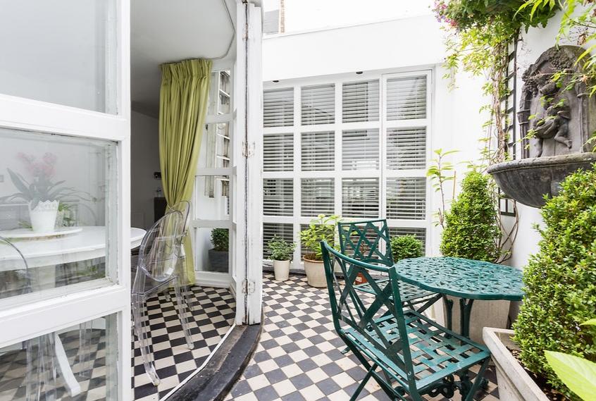 An outdoor patio area features a checkered tiled floor, accompanied by a green table and two matching chairs. Lush potted plants surround the space, while large windows with shutters allow natural light to filter in. Decorative elements include a water feature and greenery along the walls.