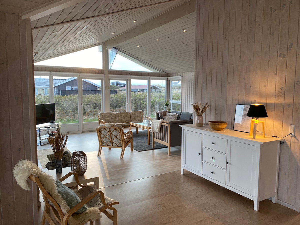 A bright living area features large windows that invite natural light, offering views of the surrounding landscape. Comfortable seating arrangements include a sofa and wicker chairs, complemented by a simple side table and decor elements that enhance the serene ambiance.