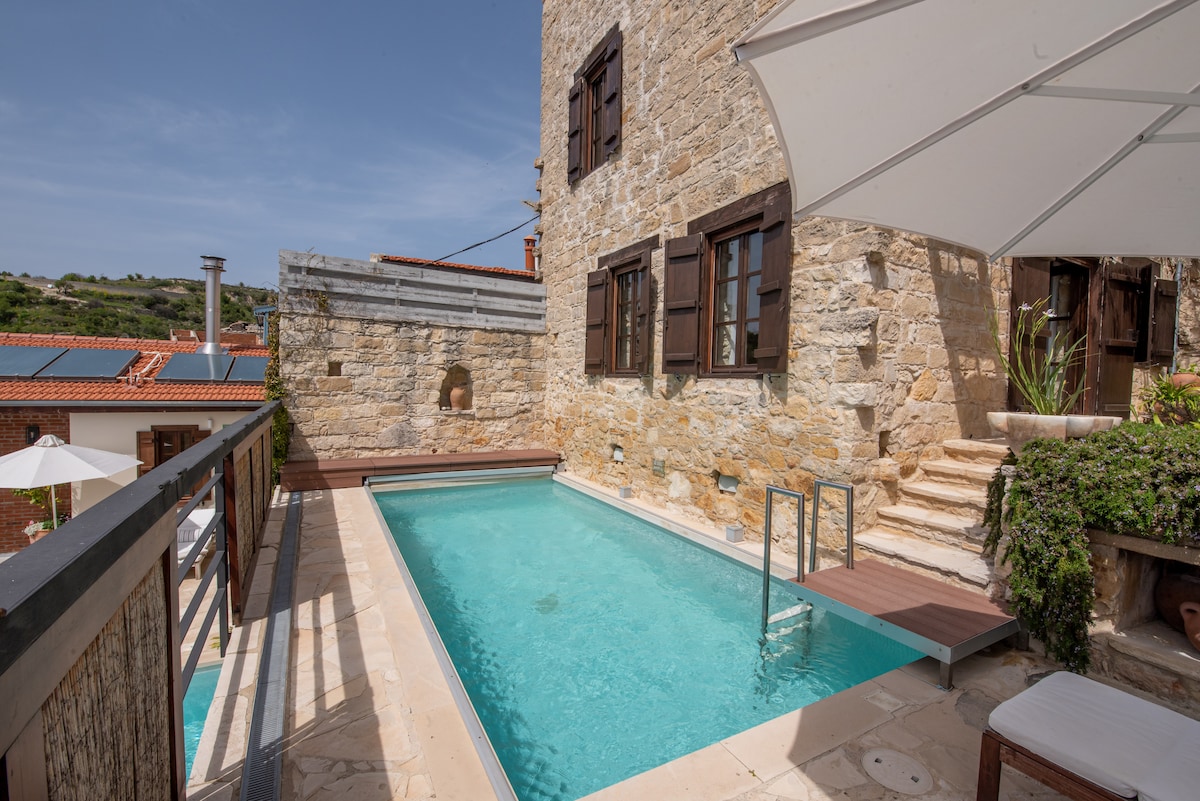 A private pool is framed by natural stone walls, providing a refreshing escape. Sun loungers and an umbrella offer comfortable seating beside the pool. Large windows allow natural light to flood the space, enhancing the serene outdoor atmosphere.