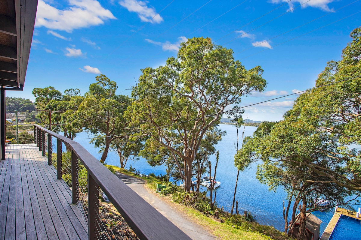 A wooden deck extends outward, offering scenic views of the tranquil bay and surrounding trees. The natural landscape features a clear blue sky and hints of watercraft visible in the distance, creating a serene outdoor space ideal for relaxation.