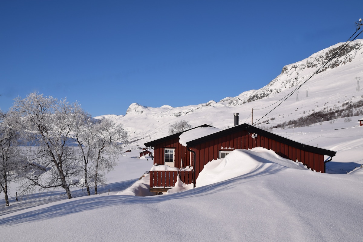 A red cabin is nestled in a snowy landscape, surrounded by gentle slopes and frost-covered trees. The expansive view includes distant mountains under a clear blue sky, creating a serene winter setting. Snow drifts gather around the cabin, emphasizing its secluded location.