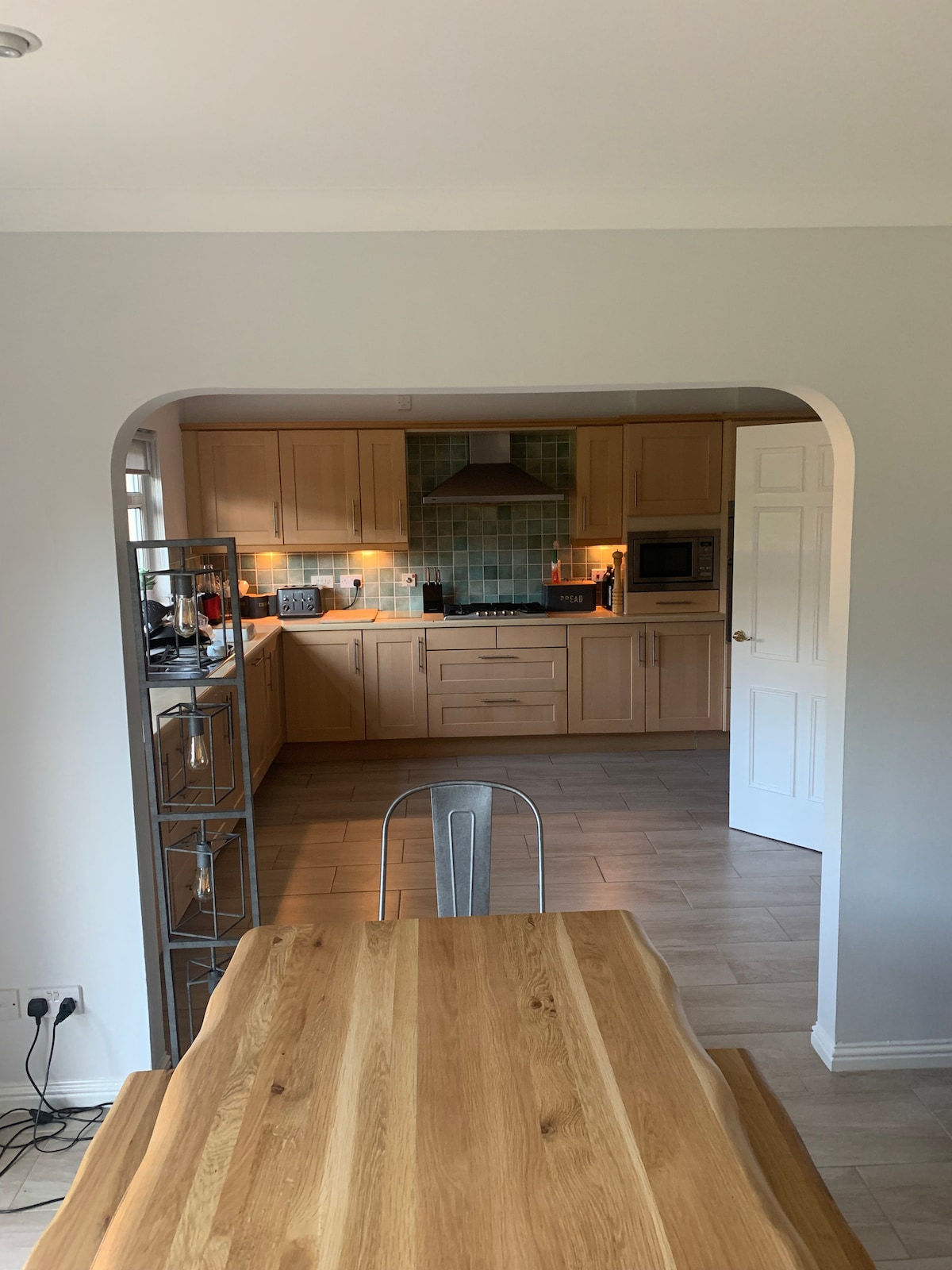 A well-equipped kitchen is visible through an arched entryway, featuring wooden cabinets and a modern range hood. A dining table made of light wood is set beneath a single metal chair, with soft lighting highlighting the functional space.