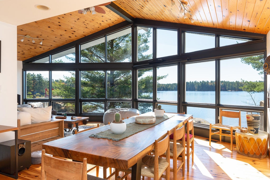A spacious dining area with a solid wood table seats ten, featuring large windows that offer views of Kahshe Lake. Natural light fills the room, highlighting the vaulted ceiling and wooden accents throughout the space.