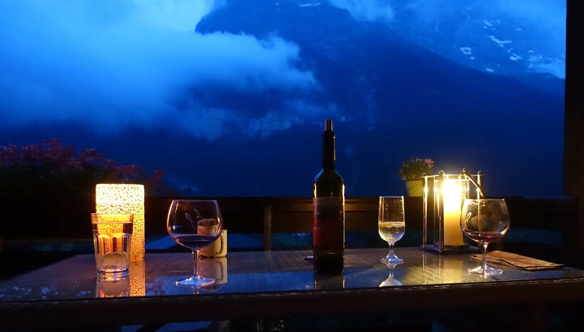An outdoor table is set with glasses and a wine bottle, surrounded by softly glowing candles. The backdrop features a mountain landscape beneath a twilight sky, enhancing the serene ambiance. Flowers can be seen along the edge, adding a touch of nature.
