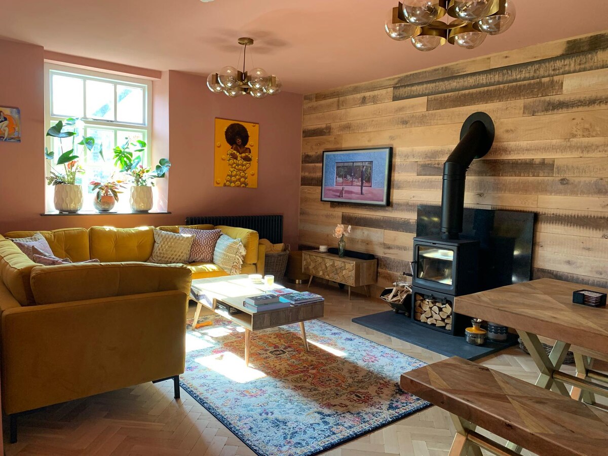 An open living area features a bright yellow sofa adorned with cushions, situated beside a wooden accent wall. A sleek black wood burner stands next to a coffee table on a patterned rug. Natural light enters through windows with greenery visible.