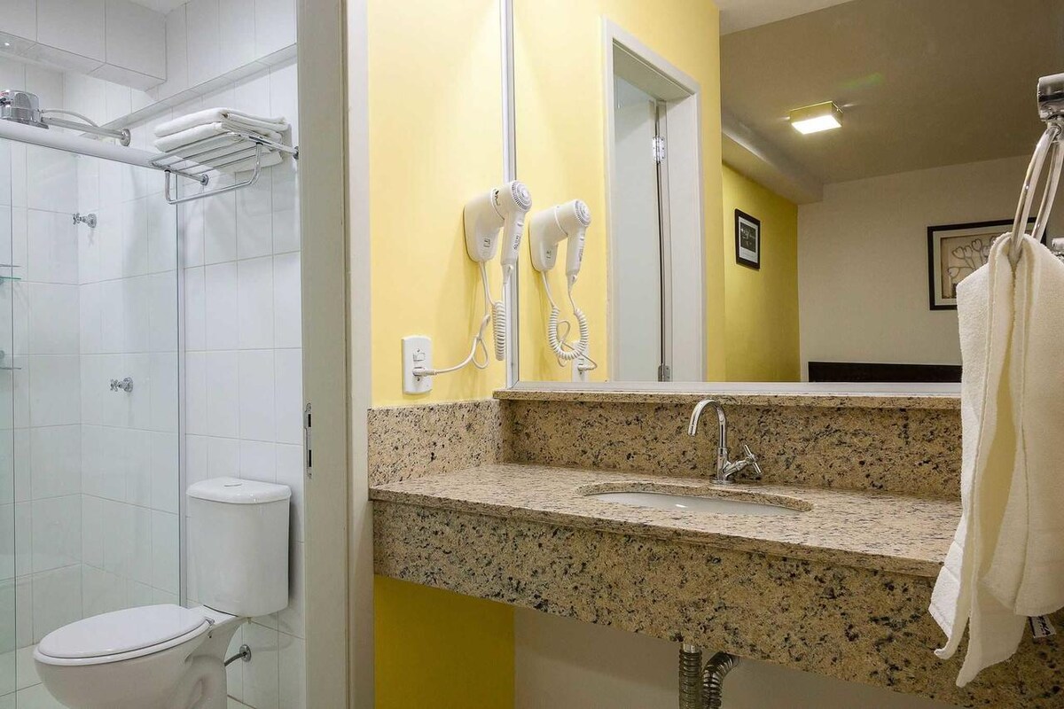 A modern bathroom features a granite counter with a sink and large mirror. White tiles cover the walls, while a shower area is partially visible. Essential toiletries, including two hairdryers on the wall, are available for guest use.
