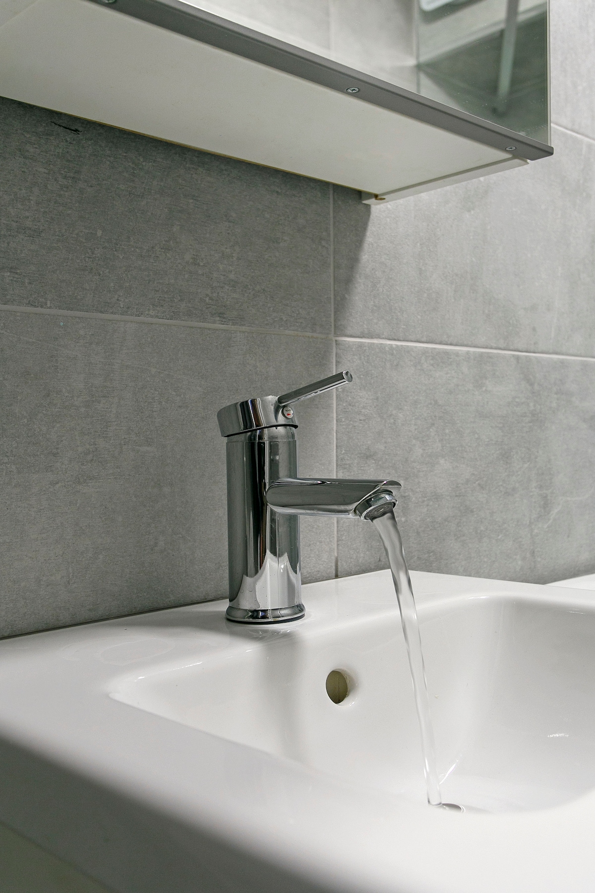 A sleek chrome faucet is shown with water flowing into a white sink. The background features gray tiles that create a modern aesthetic. A mirror is visible above, reflecting the faucet area.