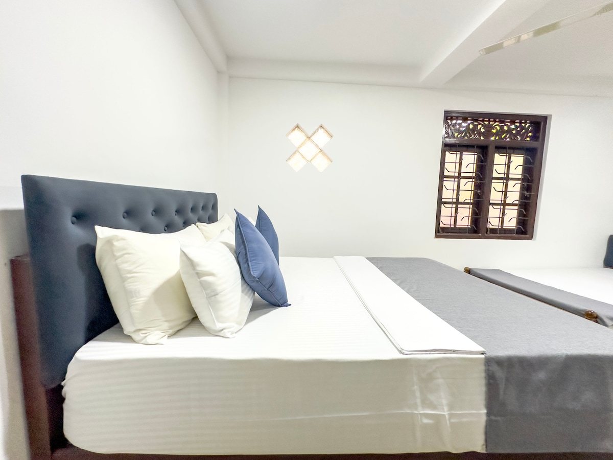 A spacious bedroom features a large bed with white and blue accent pillows. Natural light filters through a decorative window, enhancing the bright, neutral tones of the room. The minimalist design promotes a calm atmosphere, ideal for rest and relaxation.