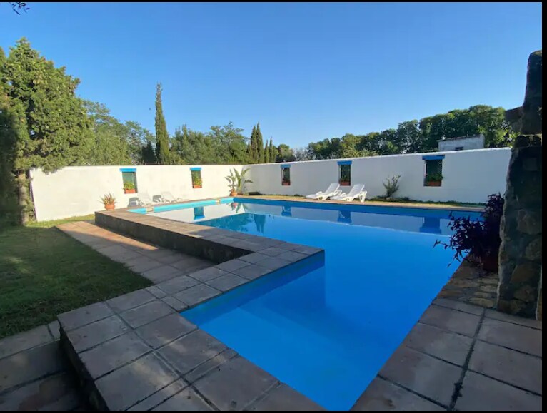 A serene swimming pool is surrounded by white walls and greenery, featuring clear blue water reflecting the sky. Sun loungers are positioned along the edge, offering a space for relaxation. The pool area is complemented by neatly trimmed grass and potted plants, enhancing the tranquil setting.