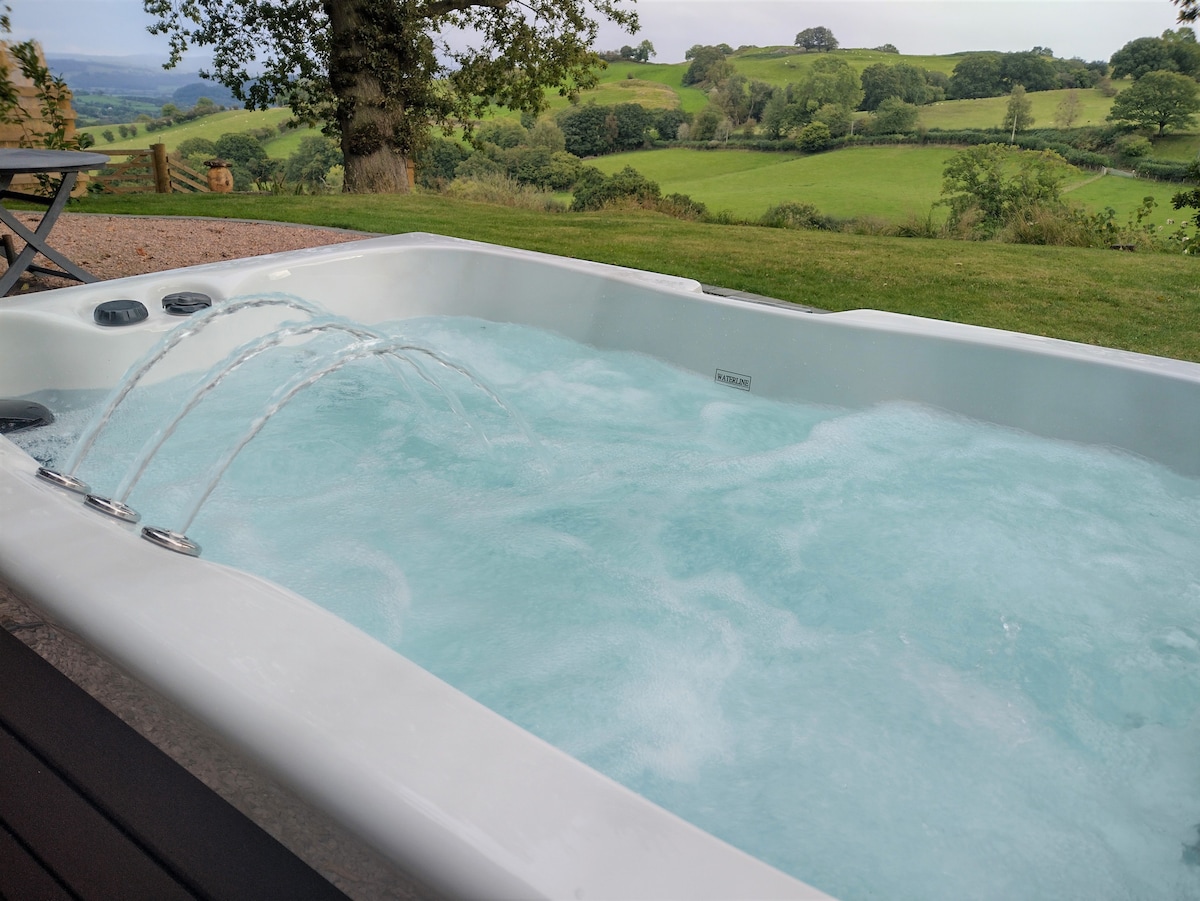 A private covered hot tub is seen, featuring water jets in action. The sparkling blue water contrasts with the green countryside and rolling hills in the background, providing a serene environment for relaxation.