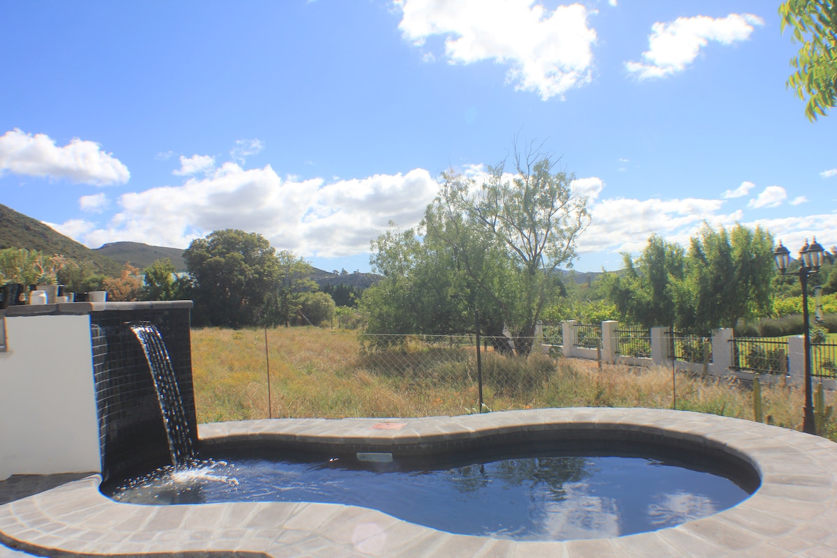 A private pool with a sleek design is surrounded by natural landscapes, showcasing green fields and distant hills. A gentle water feature cascades into the pool, reflecting the blue sky dotted with fluffy clouds, enhancing the serene outdoor setting.