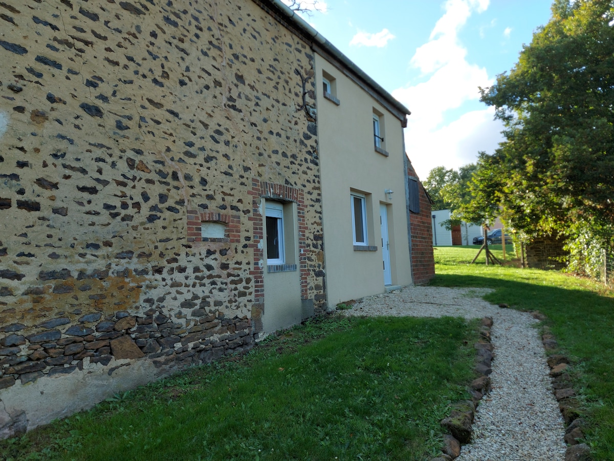 The exterior of the gîte features a combination of rustic stone and smooth wall surfaces. A gravel path leads to the main entrance, bordered by a green lawn. Surrounding trees provide a natural backdrop, enhancing the serene ambiance of the countryside setting.