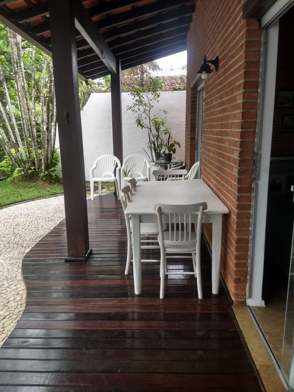 An outdoor patio area is shown, featuring a white table surrounded by four chairs. The wood flooring complements the natural surroundings, with greenery visible in the background. The space is covered by a wooden roof, providing a shaded area for relaxation.