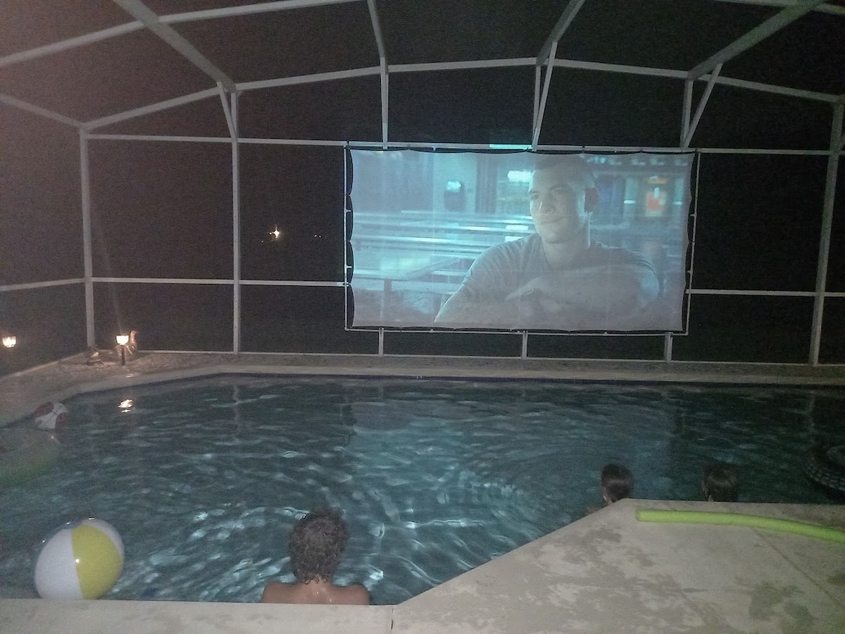 An outdoor movie setup is showcased beside a private swimming pool. A large screen is illuminated with a film, while poolside lights cast a soft glow. A beach ball and pool float are visible, enhancing the relaxed atmosphere.