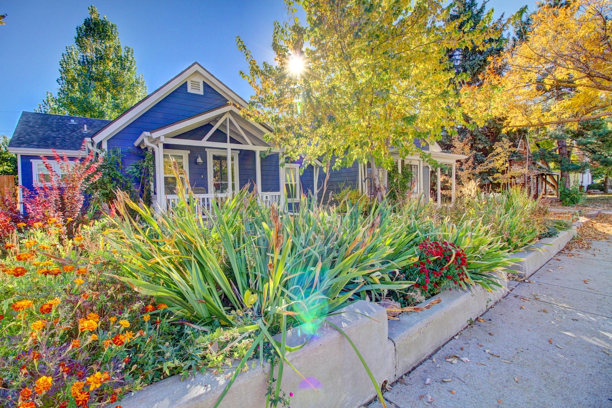 A charming bungalow is framed by a vibrant garden filled with blooming flowers and lush greenery. Sunlight filters through the trees, creating a bright atmosphere for the welcoming porch with white rocking chairs. The pathway leads up to the home, inviting relaxation and enjoyment of the outdoors.