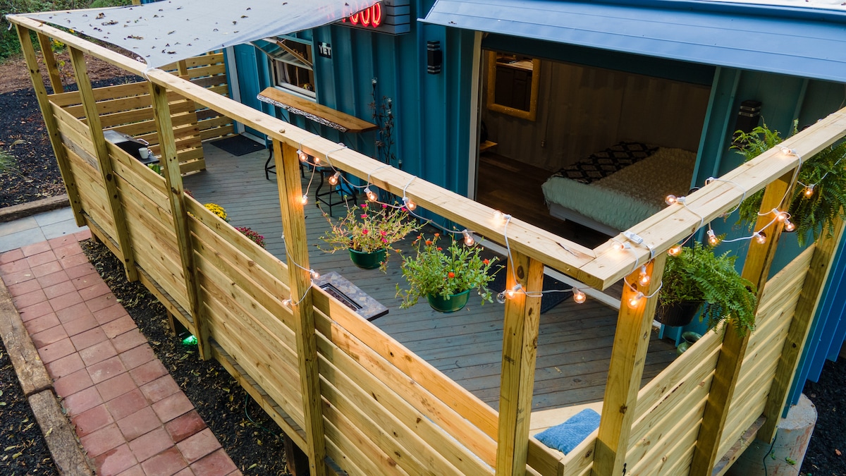 An enclosed deck area is highlighted with warm string lights and potted plants. Wooden railings create a secure space, while a glimpse of the interior shows a cozy sleeping area. The deck features a shaded section, offering comfort for outdoor relaxation.