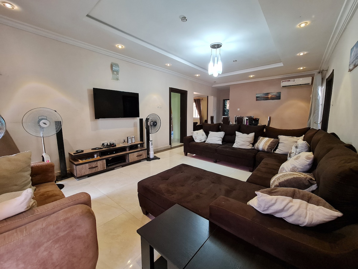 A spacious living area is highlighted by a large sectional sofa adorned with various cushions. A flat-screen television is mounted on the wall, and a coffee table sits in the center. Two standing fans contribute to a comfortable environment, complemented by soft lighting from ceiling fixtures.