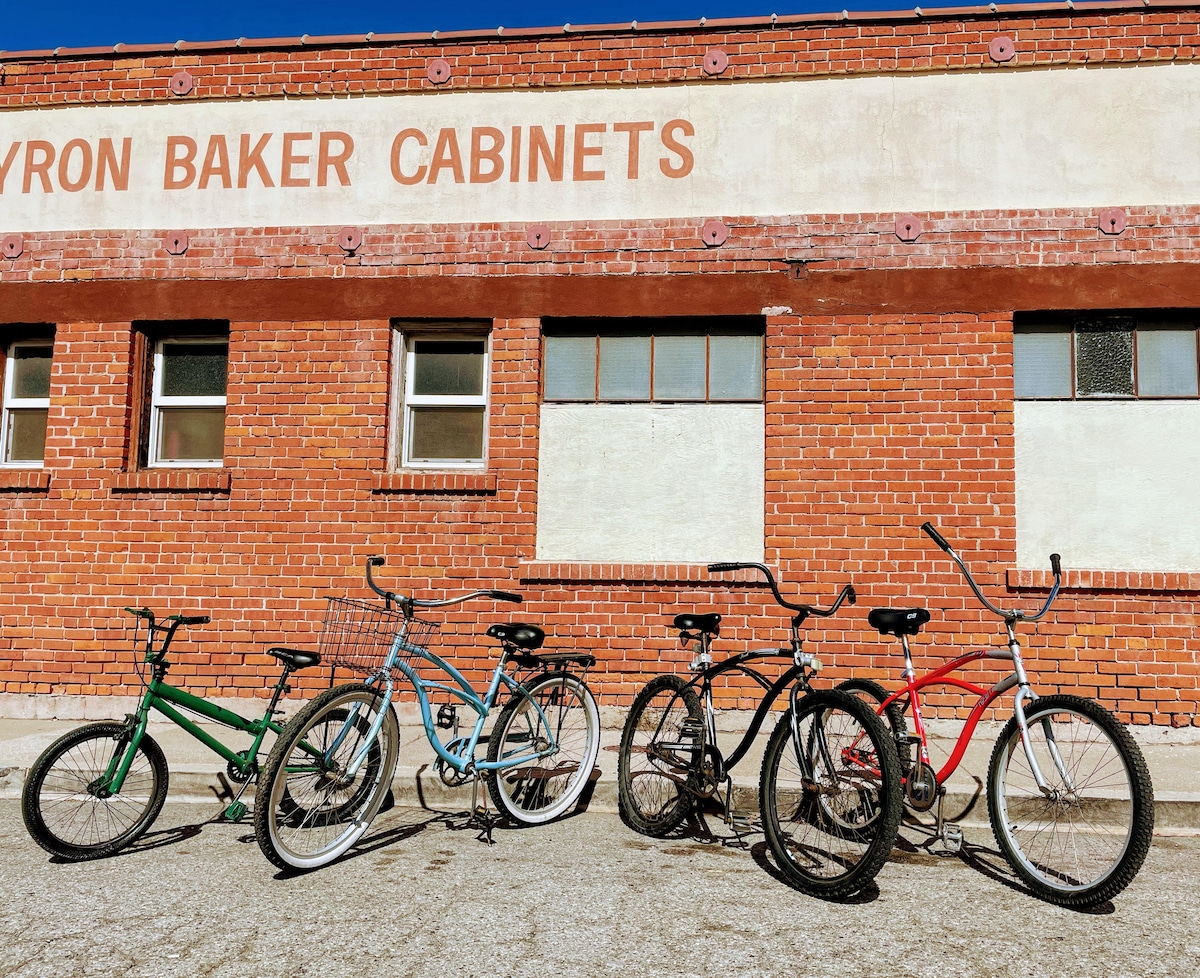 A collection of six beach cruiser bikes in varying colors, including green, blue, black, and red, is lined up next to a red-brick building. The bikes display traditional designs with comfortable seats and wide handlebars, ideal for leisurely rides around Ventura.