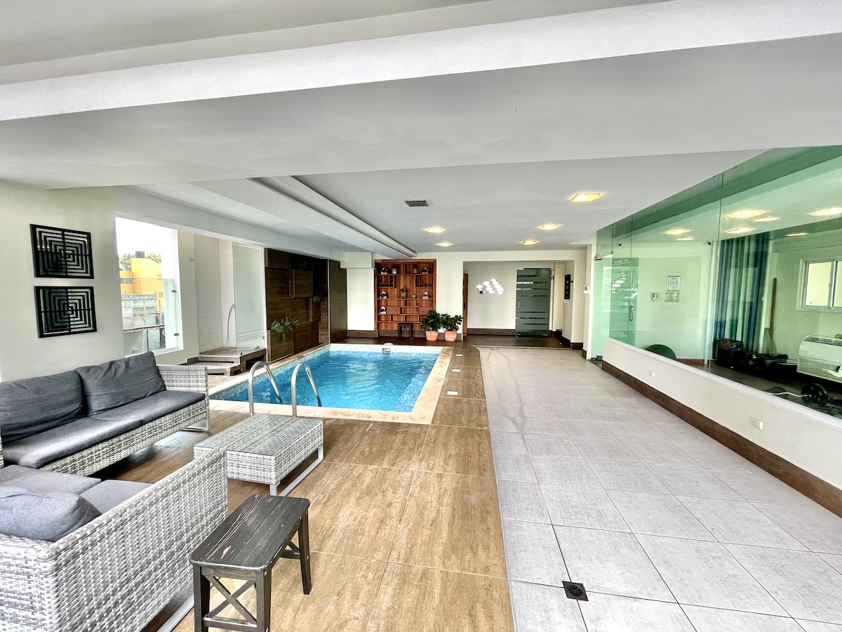An indoor area features a swimming pool surrounded by contemporary furniture, including a sectional sofa and a small table. Natural light enters through large windows, and decorative accents are visible along the wall. The space is designed for relaxation and leisure.