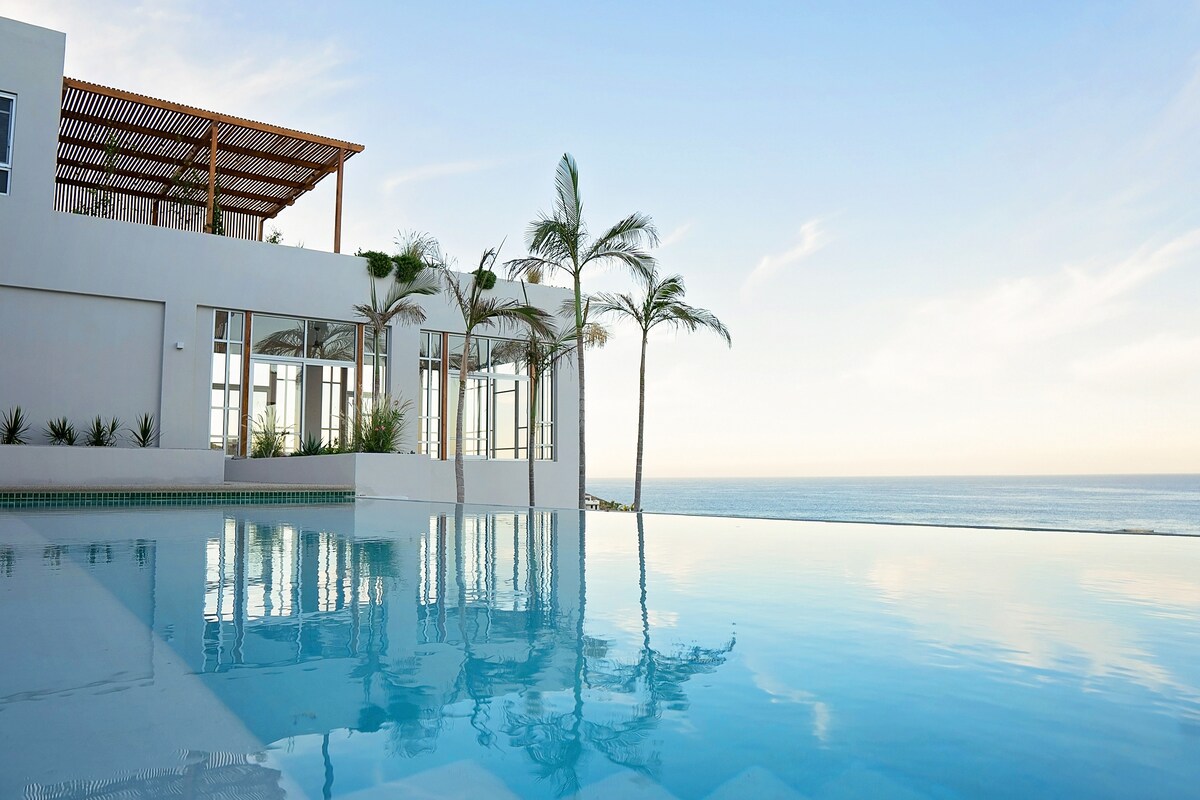 An infinity pool reflects the clear blue sky and surrounding landscape, seamlessly merging with the ocean in the distance. Palm trees are visible, swaying gently beside the pool while large windows of the building showcase a bright and airy interior.