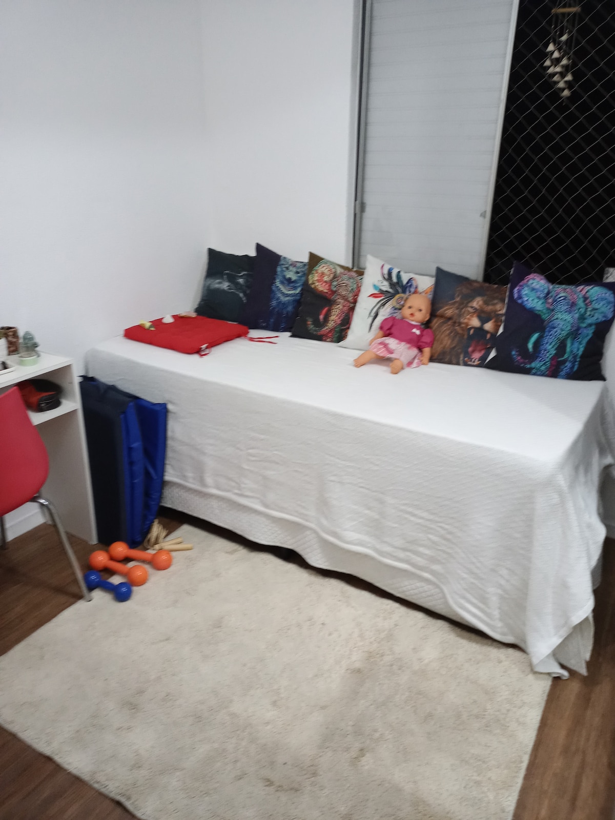 A cozy space features a daybed covered in a white blanket, surrounded by colorful cushions with animal motifs. A small, bright red chair is positioned nearby, and a soft rug is placed on a wooden floor. Fitness weights rest on the floor, adding utility to the area.