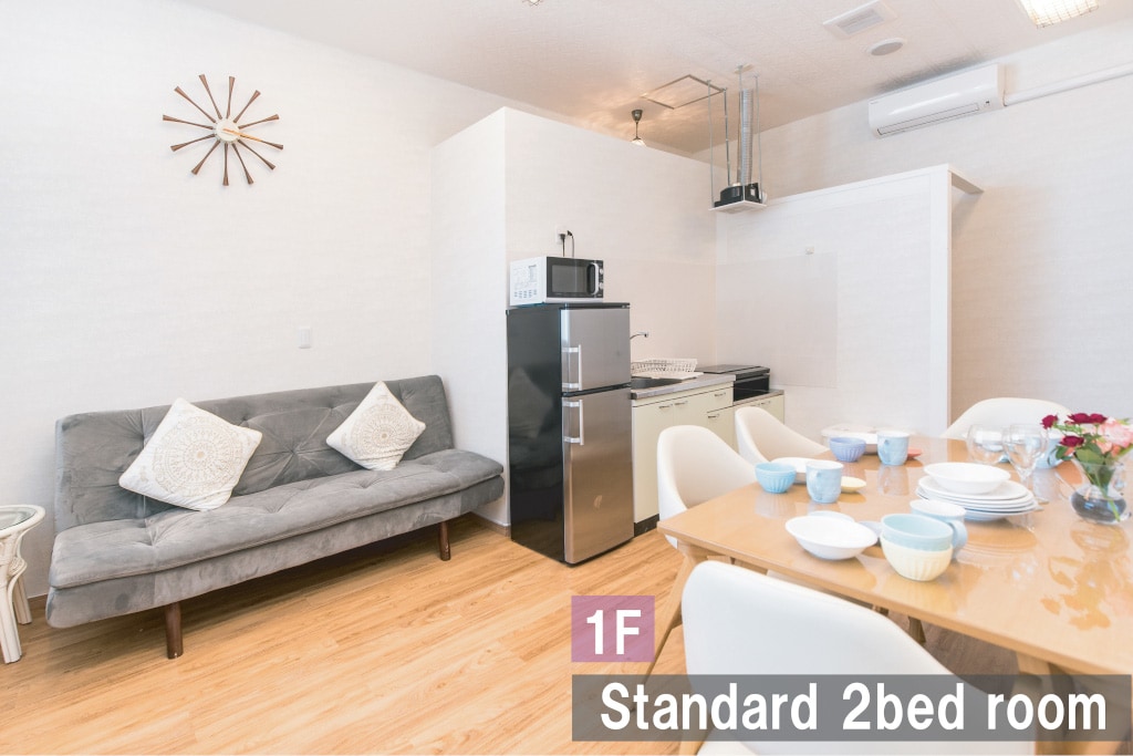 A standard two-bed room is depicted, featuring a comfortable gray sofa with decorative cushions. A dining table is set with dishes and utensils, ready for meals. A compact kitchenette, equipped with a microwave and stovetop, is visible in the background, complemented by light hardwood flooring.