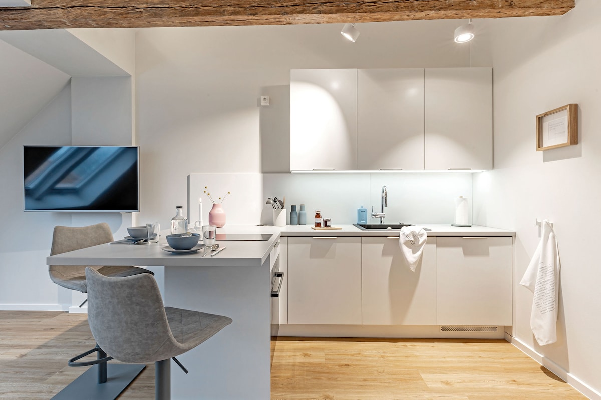 A modern kitchen area features sleek white cabinetry and a minimalist design. A compact dining table with two upholstered chairs is visible, along with a subtle, illuminated backsplash that complements the light wooden floors and exposed beams overhead.