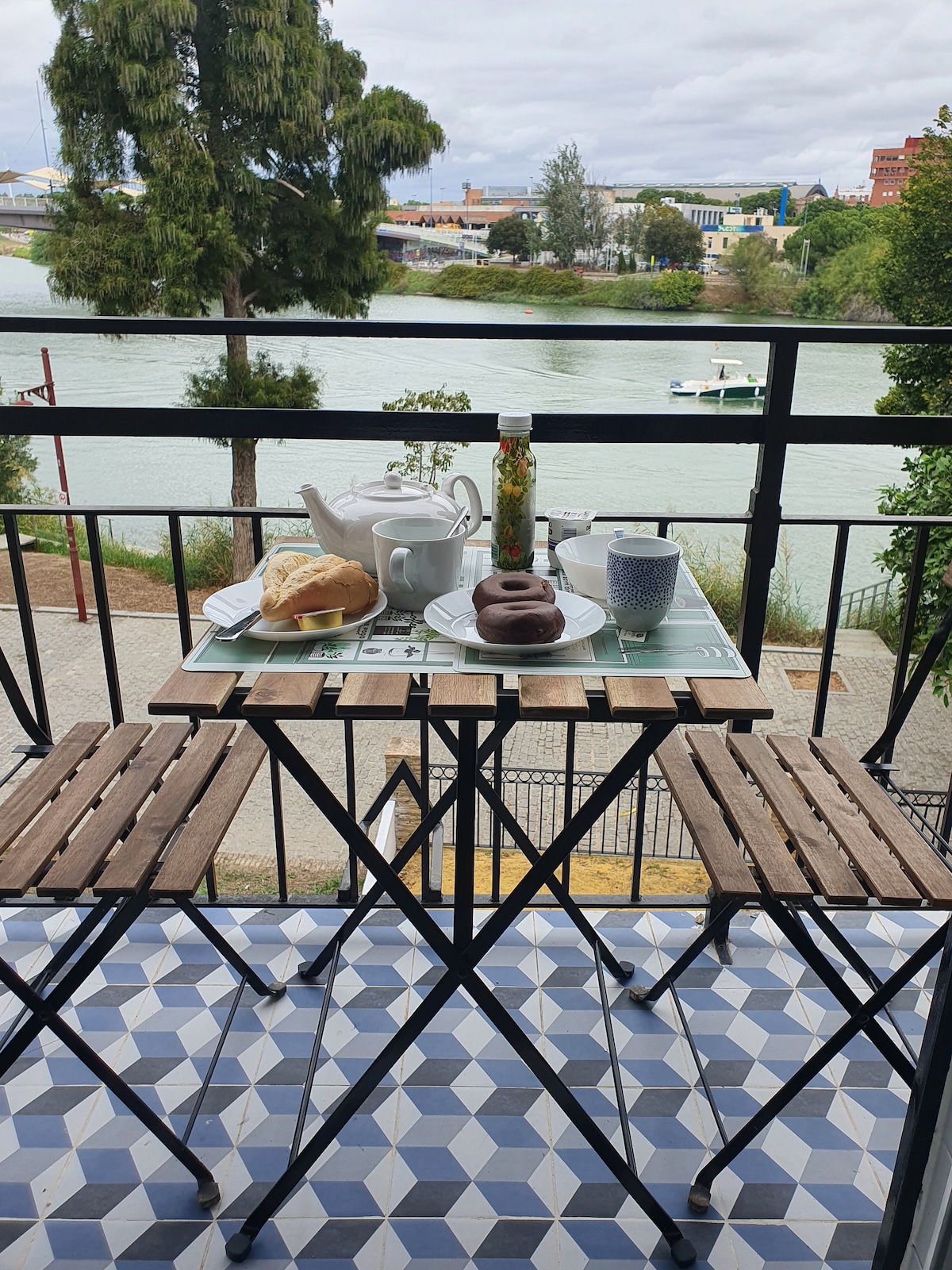 A small balcony table is set for breakfast, featuring a teapot, cups, pastries, and a decorative bottle. The table is surrounded by two wooden chairs, with a view of the river and greenery beyond, creating a serene dining space.