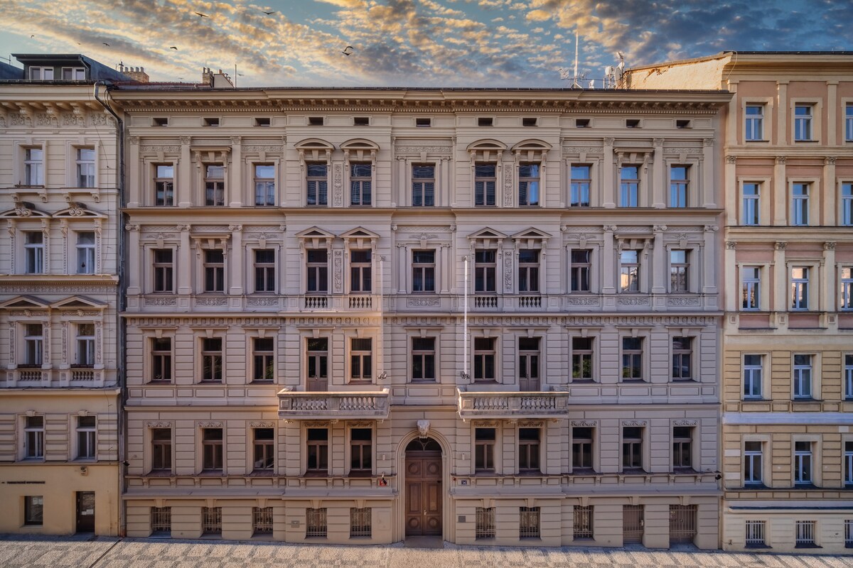 The historic building presents a classic façade, characterized by elegant architectural details and multiple windows. Balconies embellish the exterior, providing visual interest. The soft morning light enhances the intricate designs, while the sky is adorned with wispy clouds, creating a serene backdrop.