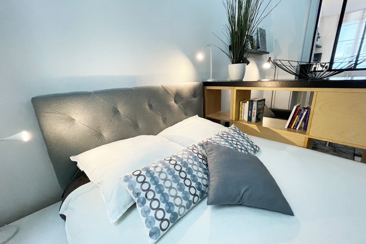 A well-arranged bedroom features a comfortable bed with an upholstered headboard and several decorative pillows. Soft lighting is provided by bedside lamps, and a stylish shelf displays books and decorative items, contributing to a cohesive modern aesthetic.