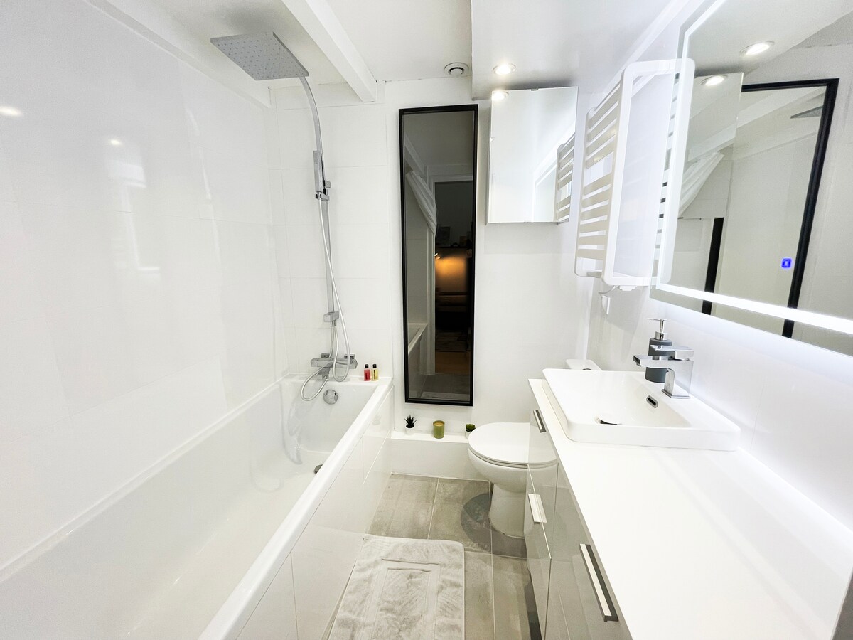 The bathroom features a bathtub with a rain shower and a large LED mirror above the vanity. A glass window allows natural light, while modern fixtures, including an electric towel dryer, add functionality. The clean, minimalist design is accented by light-colored tiles.