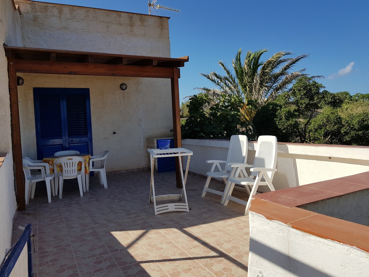 A spacious terrace is presented, featuring a shaded area supported by wooden beams. Four white chairs surround a circular table, while two sun loungers are positioned for relaxation. Lush greenery is visible in the background, complemented by a clear blue sky.